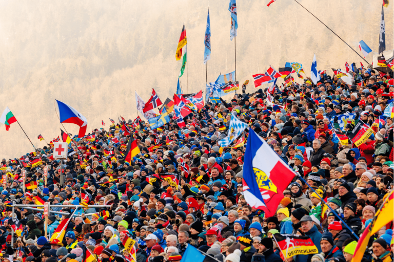 Nahaufnahme der dicht gedrängten Zuschauertribüne beim Biathlon Weltcup. Ein Meer aus Fans in bunter Winterkleidung schwenkt Flaggen verschiedener Nationen (u.a. Deutschland, Norwegen, Frankreich, Tschechien), was die internationale Begeisterung des Events verdeutlicht.