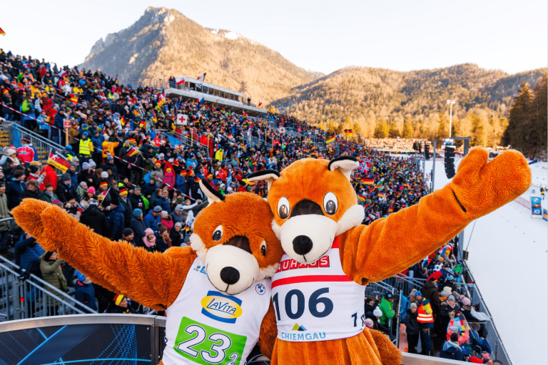 Die beiden Biathlon-Maskottchen (Füchse) posieren gut gelaunt mit ausgebreiteten Armen vor der vollbesetzten Tribüne in Ruhpolding. Im Hintergrund erstreckt sich die beeindruckende Bergkulisse unter blauem Himmel.