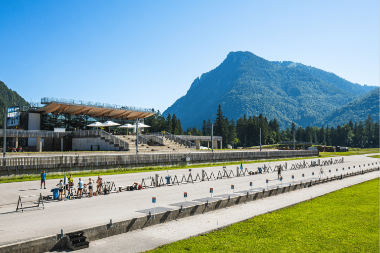 Die Chiemgau Arena im Sommerbetrieb. Sportler trainieren am Schießstand auf Rollskiern bei strahlendem Sonnenschein. Das moderne Stadiongebäude und der markante Berggipfel im Hintergrund unterstreichen die Nutzung als Ganzjahres-Stützpunkt.