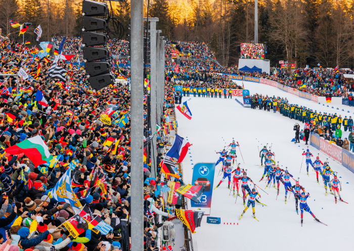 Weitwinkelaufnahme des Stadions in Ruhpolding während eines Massenstarts. Eine große Gruppe von Biathleten schiebt sich aus dem Startbereich auf die Loipe, flankiert von dicht besetzten Tribünen voller Fans, die zahlreiche internationale Flaggen schwenken.