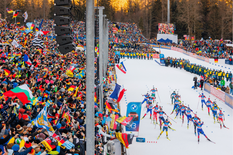 Weitwinkelaufnahme des Stadions in Ruhpolding während eines Massenstarts. Eine große Gruppe von Biathleten schiebt sich aus dem Startbereich auf die Loipe, flankiert von dicht besetzten Tribünen voller Fans, die zahlreiche internationale Flaggen schwenken.