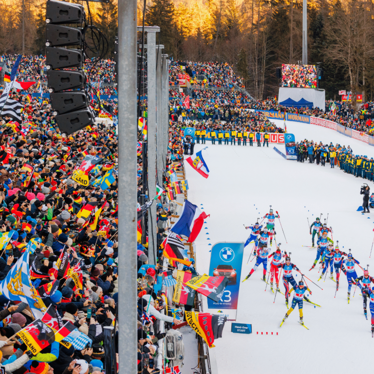 Weitwinkelaufnahme des Stadions in Ruhpolding während eines Massenstarts. Eine große Gruppe von Biathleten schiebt sich aus dem Startbereich auf die Loipe, flankiert von dicht besetzten Tribünen voller Fans, die zahlreiche internationale Flaggen schwenken.