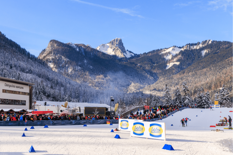 Blick auf das verschneite Stadiongelände der Chiemgau Arena vor einer markanten, schneebedeckten Bergspitze. Im Vordergrund sind die Loipe mit Werbebanden und Zuschauerbereiche zu sehen, die die weitläufige Anlage in der alpinen Landschaft zeigen.